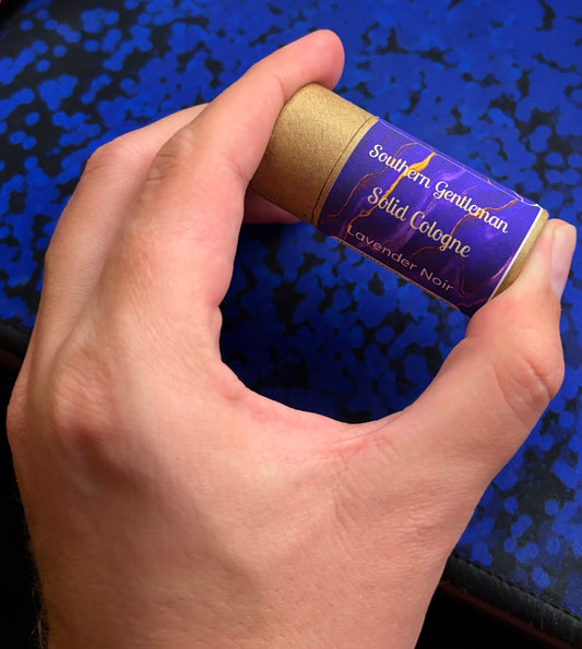 Hand holding a Southern Gentlemann Solid Cologne stick against a blue patterned background